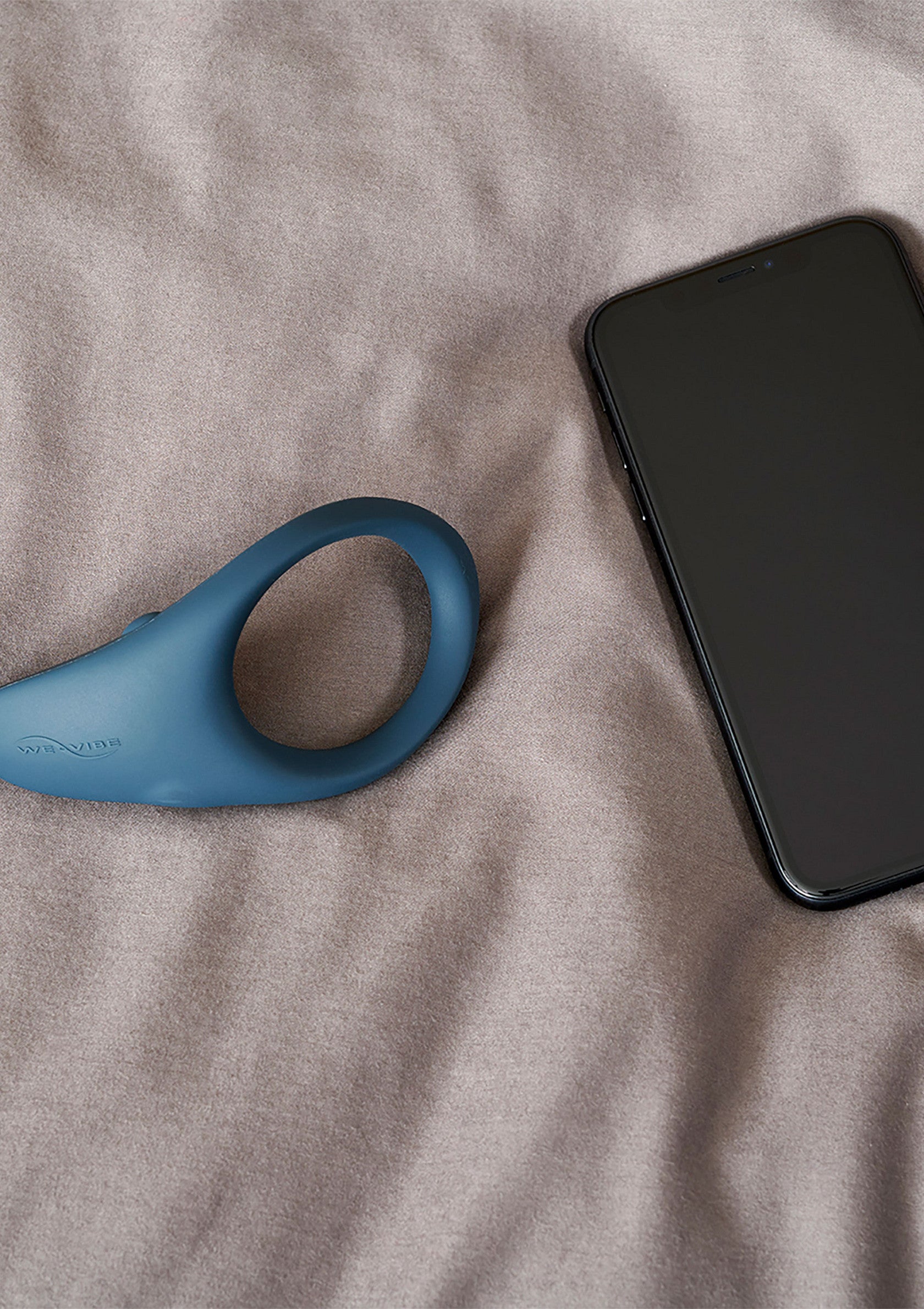 Image d'un anneau vibrant bleu à côté d'un smartphone sur un tissu froissé couleur pêche. Accessoires de bien-être sexuel et technologie moderne.; Afbeelding van een blauwe vibrerende ring naast een smartphone op een perzikkleurige, gekreukte stof. Seksuele wellness accessoires en moderne technologie.; Image of a blue vibrating ring next to a smartphone on a peach-colored, crumpled fabric. Sexual wellness accessories and modern technology.