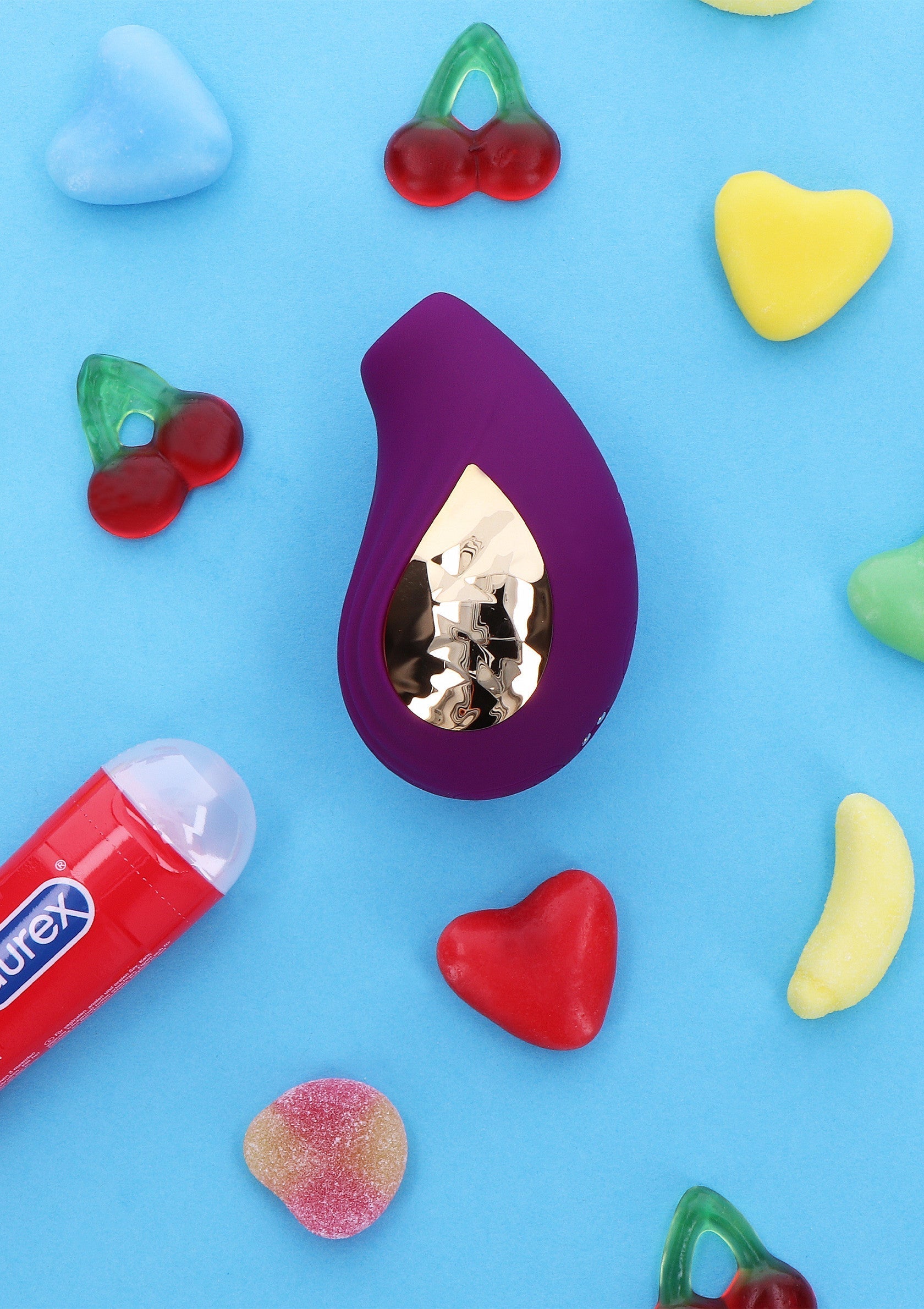 Vibrateur violet sur fond bleu avec des bonbons colorés et un tube de lubrifiant Durex.; Paarse vibrator op blauwe achtergrond met kleurrijke snoepjes en een tube Durex glijmiddel.; Purple vibrator on blue background with colorful candies and a tube of Durex lubricant.