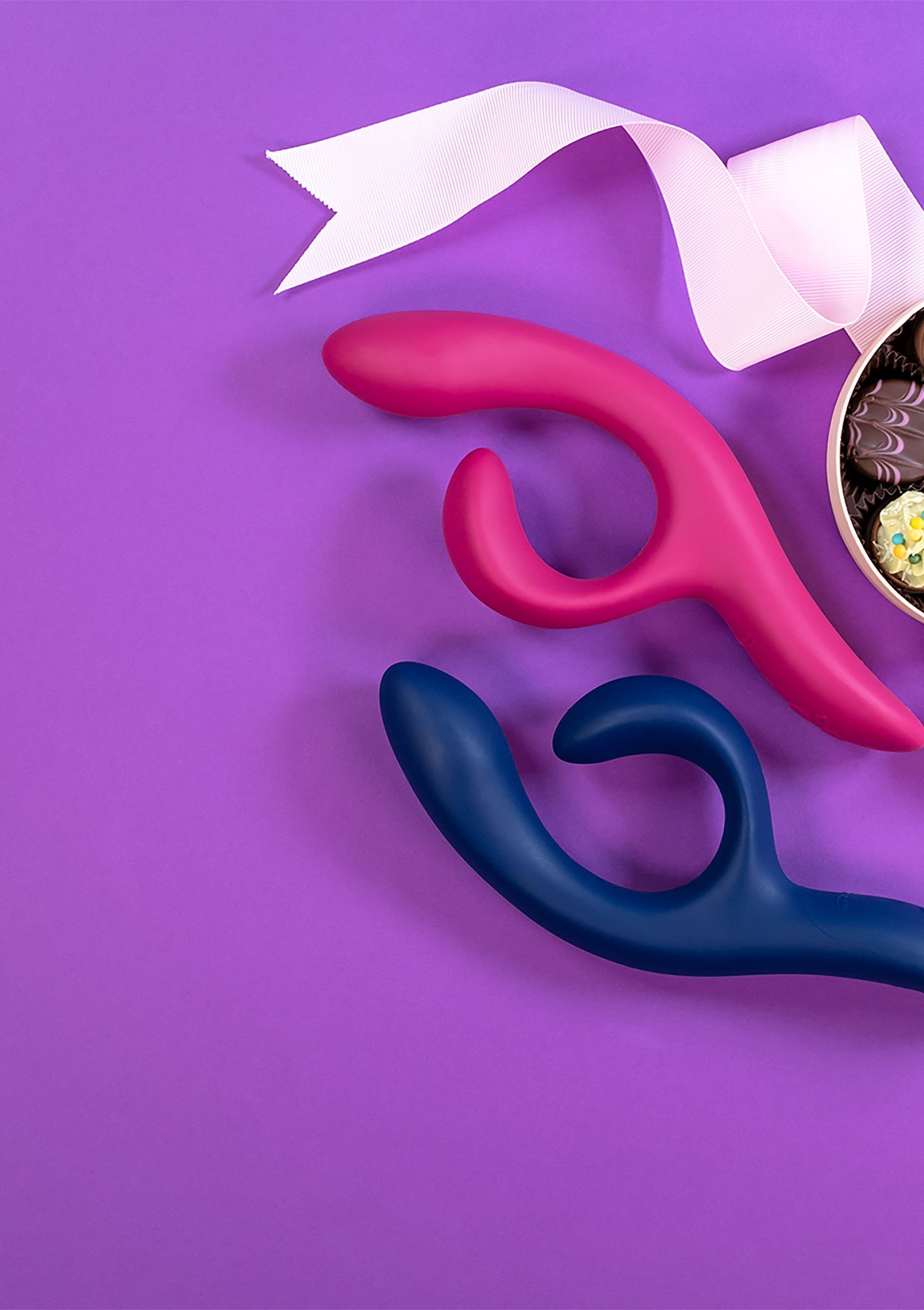 Vibrateurs roses et bleus avec ruban et chocolats sur fond violet. Cadeaux coquins pour adultes.; Roze en blauwe vibrators met lint en chocolaatjes op een paarse achtergrond. Speelse cadeaus voor volwassenen.; Pink & blue vibrators with ribbon and chocolates on purple background. Playful adult gifts.