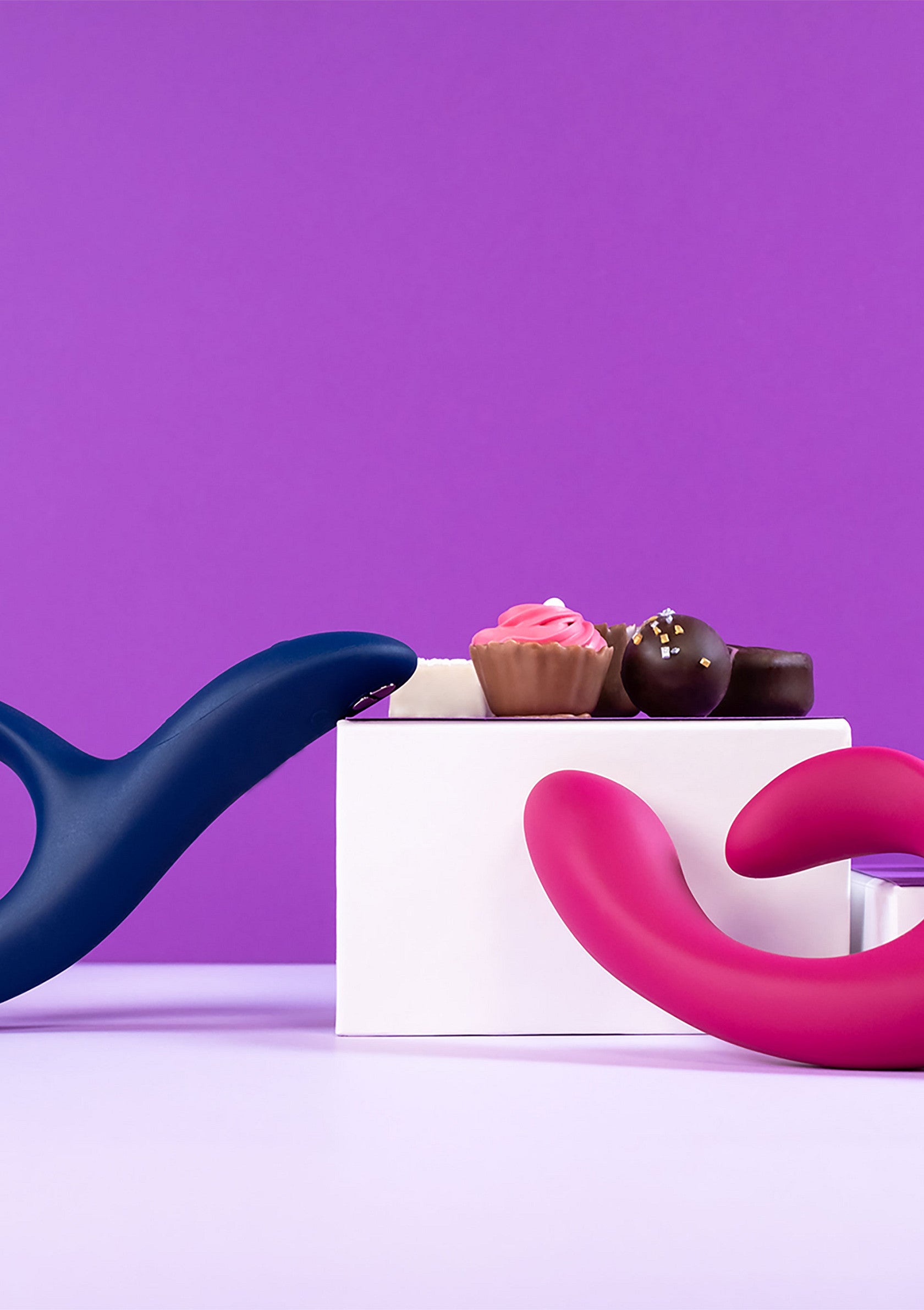 Vibromasseurs roses et bleus avec des chocolats sur fond violet. Cadeaux coquins pour adultes.; Roze en blauwe vibrators met chocolade op paarse achtergrond. Speelse cadeaus voor volwassenen.; Pink & blue vibrators with chocolates on purple background. Playful adult gifts.