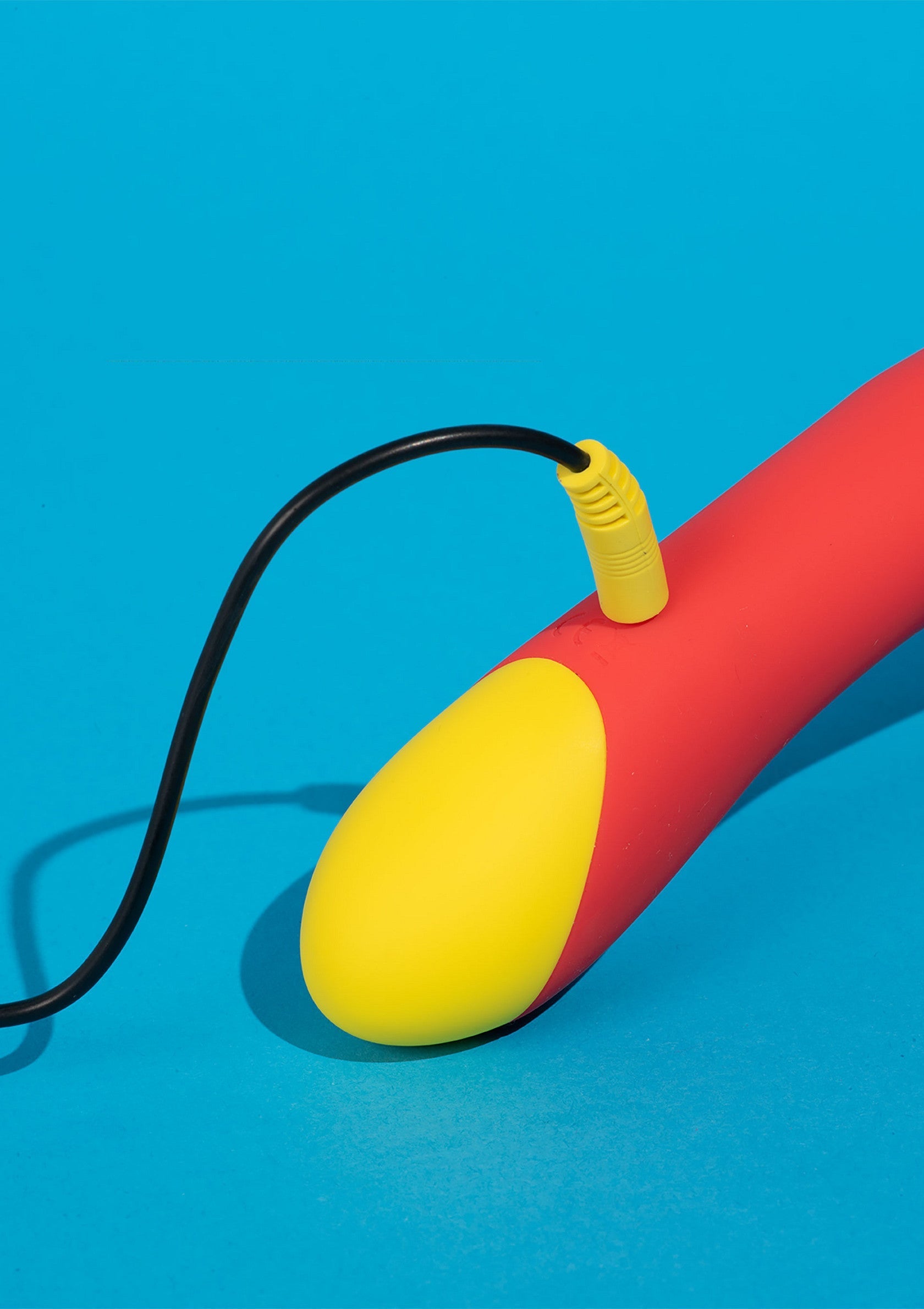 Vibromasseur rouge et jaune avec câble de chargement noir sur fond bleu.; Rode en gele vibrator met zwarte oplaadkabel op blauwe achtergrond.; Red and yellow vibrator with black charging cable on blue background.