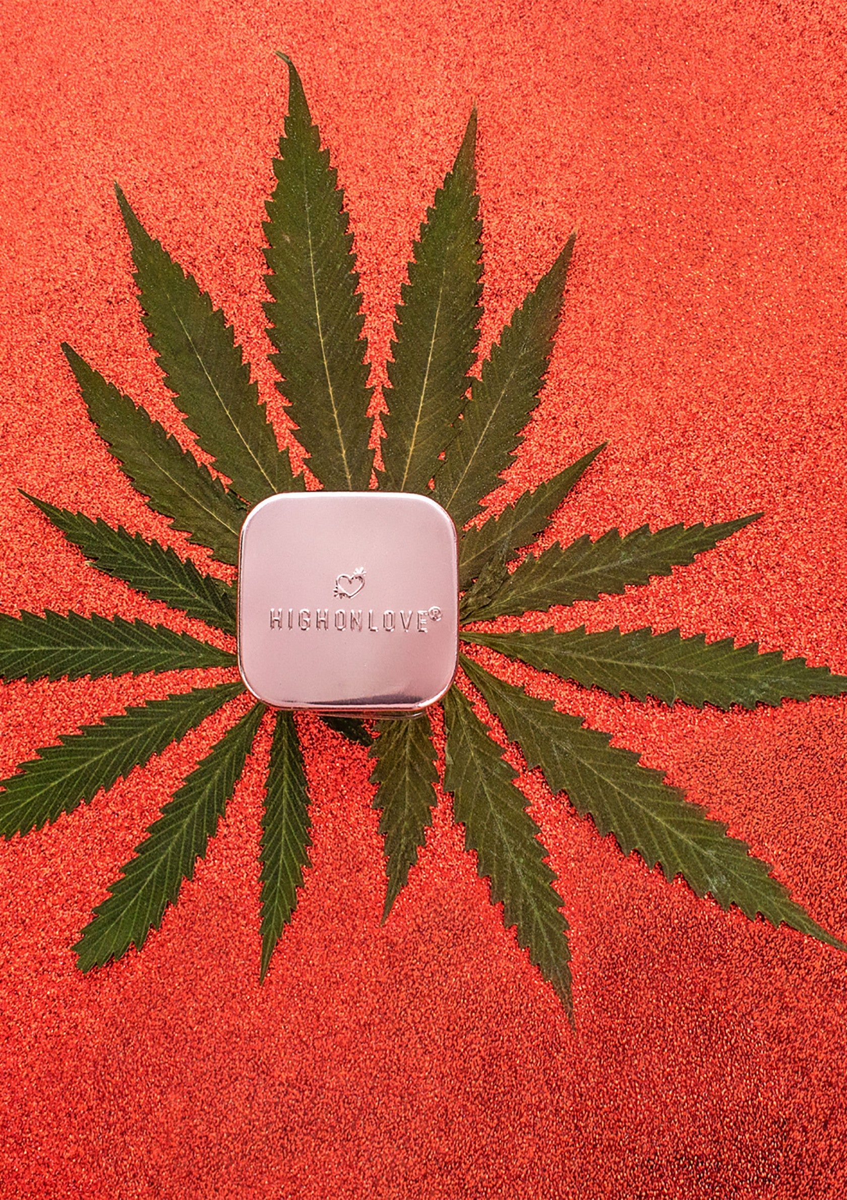 Boîte argentée 'HIGHONLOVE' sur feuille de cannabis, fond orange scintillant. Cadeau parfait et original.; Zilveren doosje 'HIGHONLOVE' op cannabisblad, fonkelende oranje achtergrond. Perfect en origineel cadeau.; Silver 'HIGHONLOVE' box on cannabis leaf, sparkling orange background. Perfect & original gift.