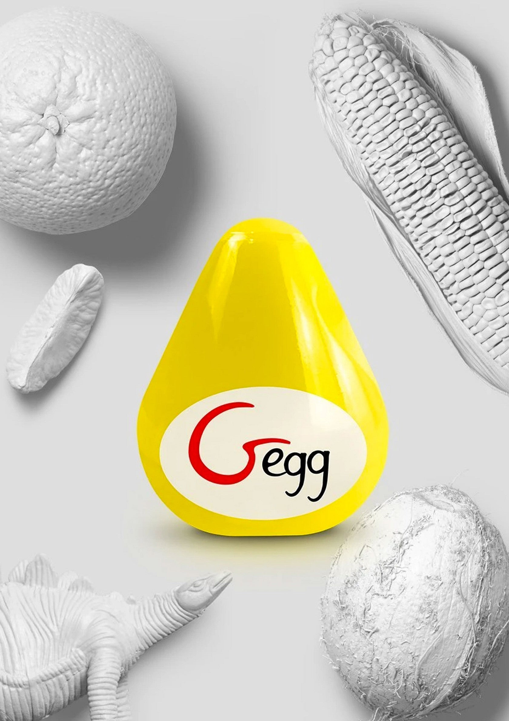 Oeuf jaune décoratif 'Gegg' entouré d'orange, maïs, noix de coco et dinosaure blancs.; Decoratief geel 'Gegg'-ei omringd door witte sinaasappel, maïs, kokosnoot en dinosaurus.; Decorative yellow 'Gegg' egg surrounded by white orange, corn, coconut, and dinosaur.
