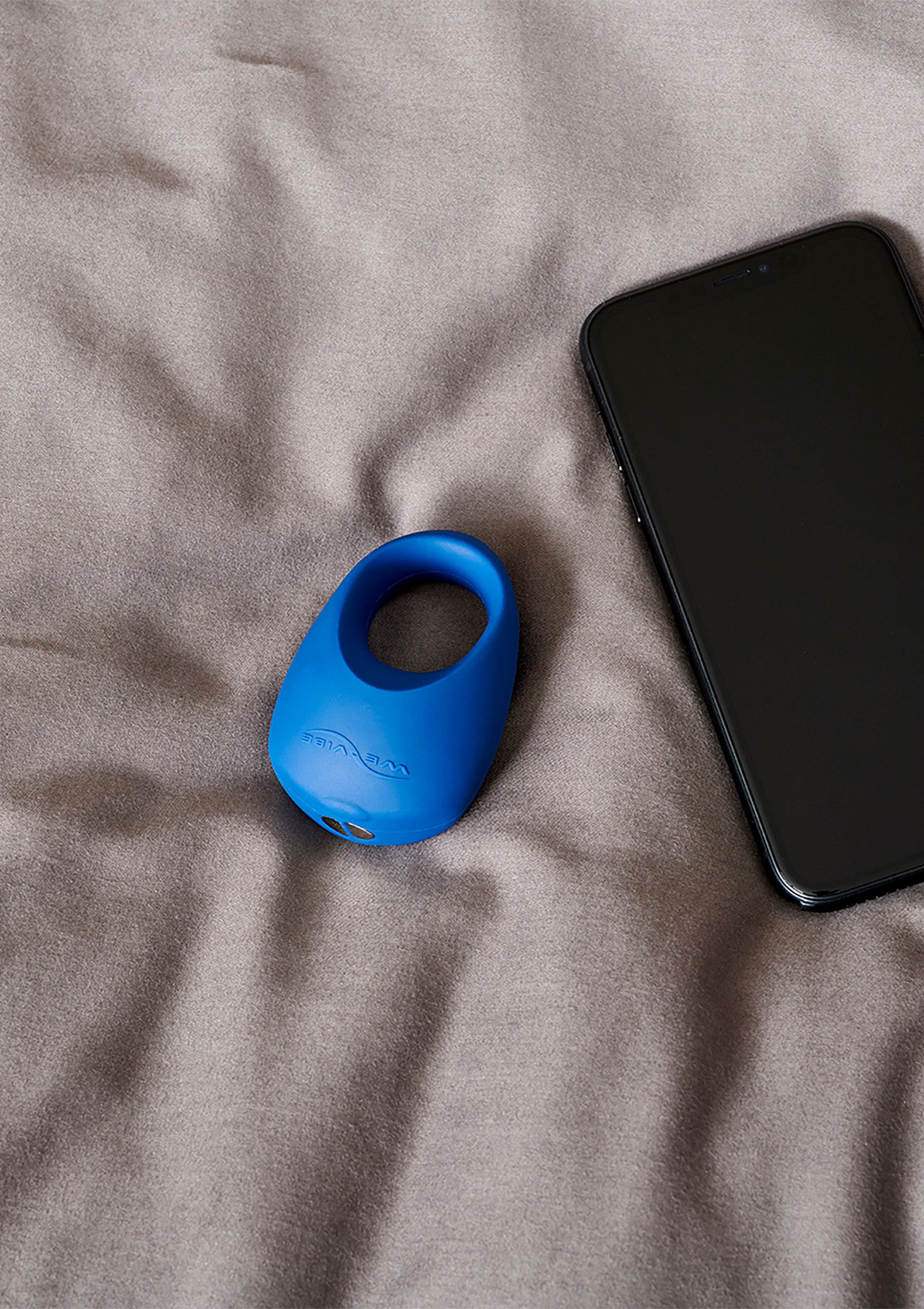 Image d'un anneau vibrant bleu pour couple, à côté d'un téléphone portable sur un drap gris.; Afbeelding van een blauwe vibrerende ring voor koppels, naast een mobiele telefoon op een grijs laken.; Image of a blue vibrating couple's ring, next to a mobile phone on a grey sheet.