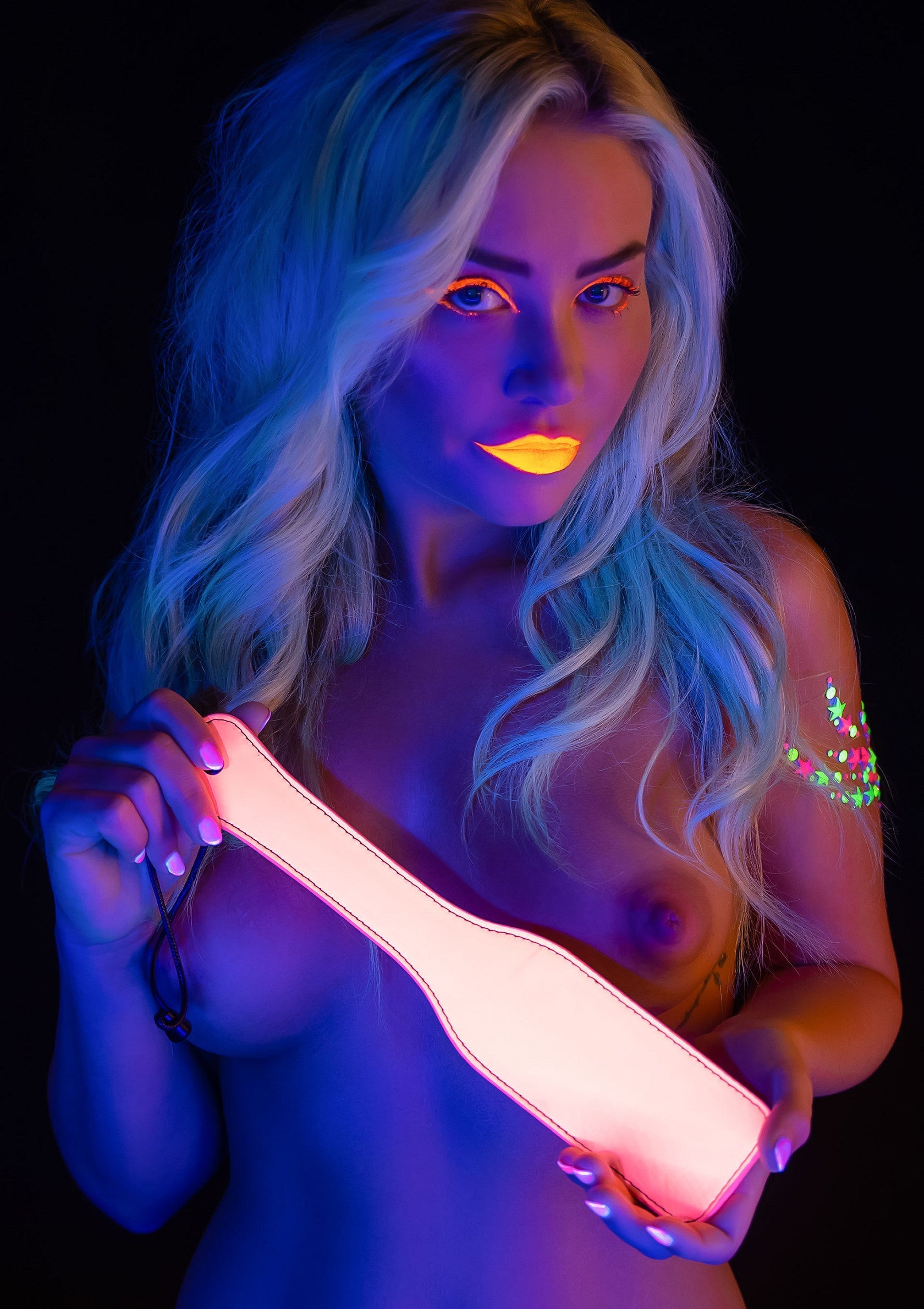 Femme sous lumière noire tenant une palette lumineuse. Accessoires BDSM et fétichistes disponibles.; Vrouw onder blacklight houdt lichtgevende paddle vast. BDSM- en fetishaccessoires verkrijgbaar.; Woman under blacklight holding glowing paddle. BDSM and fetish accessories available.