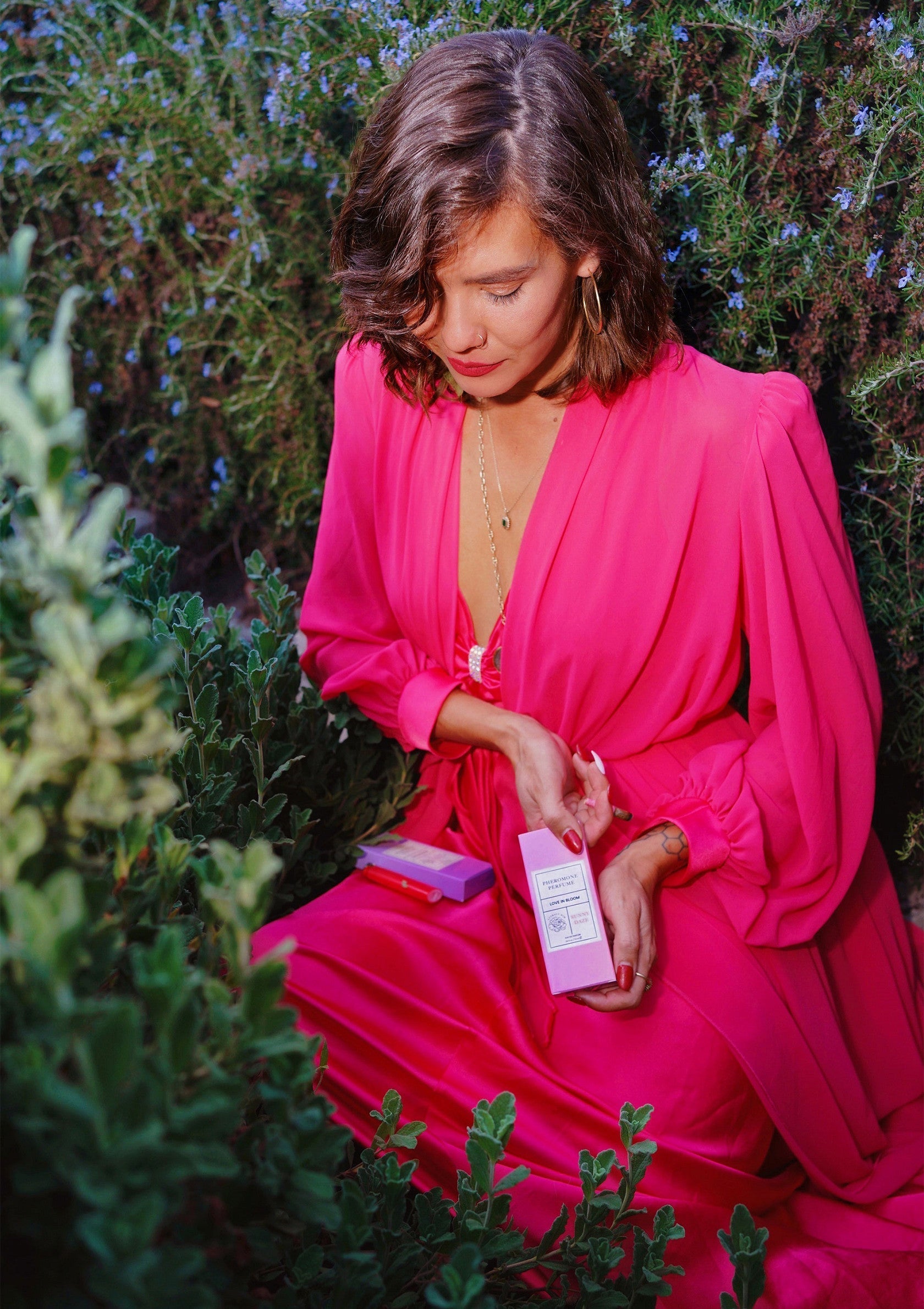 Femme en robe rose présentant un produit de beauté dans un cadre floral luxuriant. Soin de la peau et élégance.; Vrouw in roze jurk presenteert schoonheidsproduct in weelderige bloemenomgeving. Huidverzorging en elegantie.; Woman in pink dress showcasing beauty product in lush floral setting. Skincare and elegance.