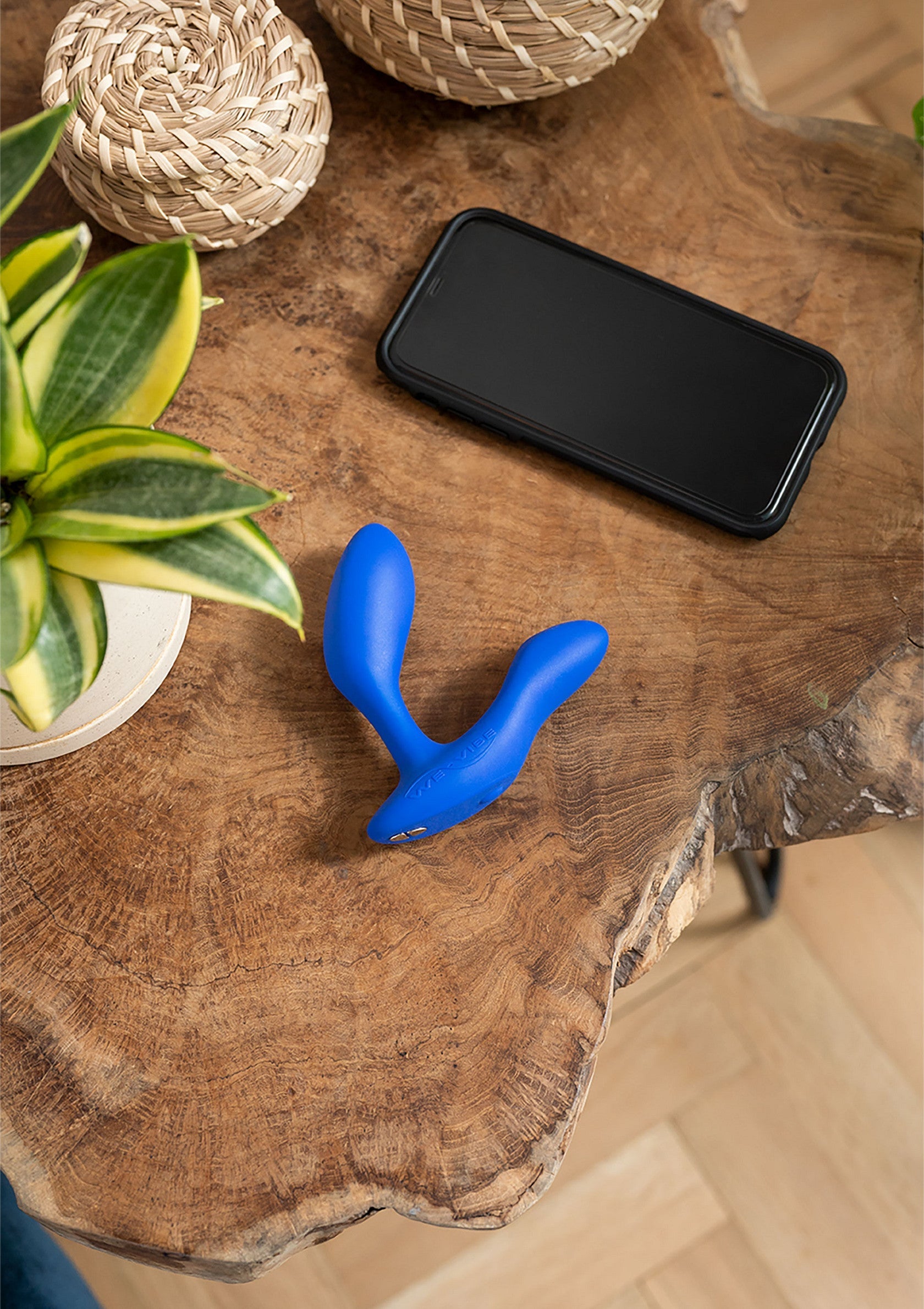 Vibromasseur bleu sur table en bois avec téléphone et plantes.; Blauwe vibrator op houten tafel met telefoon en planten.; Blue vibrator on wooden table with phone and plants.